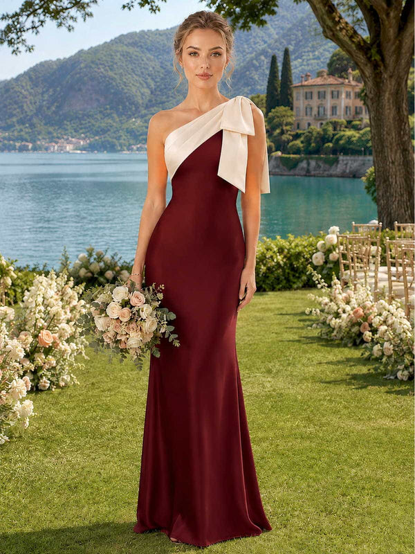 Front view of a burgundy satin floor-length bridesmaid dress with an asymmetric one-shoulder neckline, ivory contrast draped shoulder panel, oversized bow detail, fitted bodice, defined waist, and softly flared long skirt.