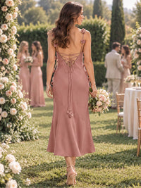 Back view of a dusty rose imitation silk tea-length bridesmaid dress featuring floral-accented spaghetti straps, a deep open back, delicate crisscross lace-up detailing, long tie ends with floral accents, fitted sheath silhouette, and softly flared hem.