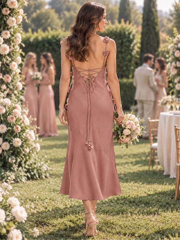 Back view of a dusty rose imitation silk tea-length bridesmaid dress featuring floral-accented spaghetti straps, a deep open back, delicate crisscross lace-up detailing, long tie ends with floral accents, fitted sheath silhouette, and softly flared hem.
