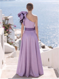 Back view of a lavender satin floor-length mother of the bride dress with asymmetric one-shoulder neckline, sculptural floral shoulder detail with black feather accents, contrasting waist sash, fitted bodice, center-back zipper, and softly flowing A-line skirt.