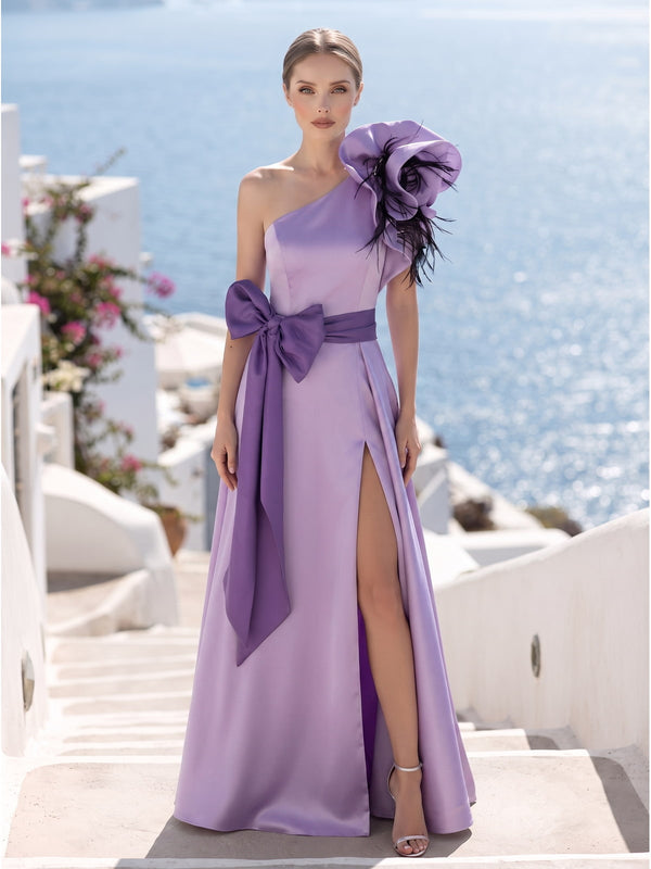 Front view of a lavender satin floor-length mother of the bride dress with asymmetric one-shoulder neckline, oversized sculptural floral shoulder detail with black feather accents, contrasting satin waist sash, oversized side bow, front slit, fitted bodice, and flowing A-line skirt.