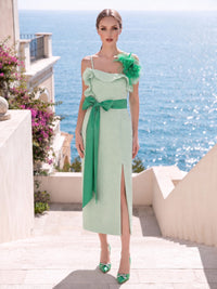 Front view of a mint green satin midi mother of the bride dress with square neckline, spaghetti straps, asymmetric layered ruffle neckline, green feather shoulder detail, contrasting green satin bow waist sash, fitted column silhouette, and front slit.