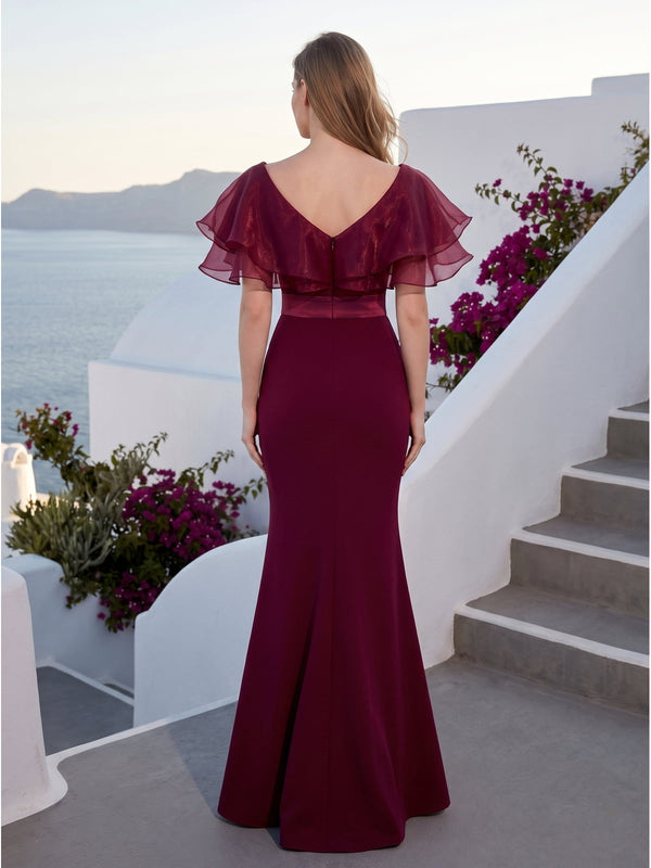 Back view of a burgundy satin floor-length mother of the bride dress with V-back neckline, layered organza ruffle cape sleeves, satin waistband, center-back zipper, fitted silhouette, and softly flared mermaid hem.