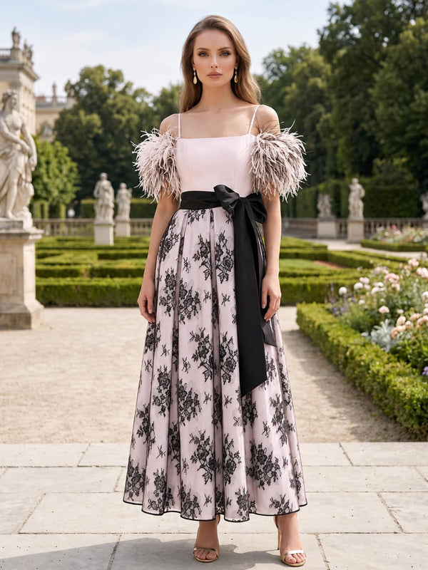 Front view of a tea-length mother of the bride dress featuring a satin square-neck bodice, delicate spaghetti straps, feather-trimmed off-shoulder sleeve accents, a contrasting satin waistband with a side bow, and a floral lace A-line skirt.