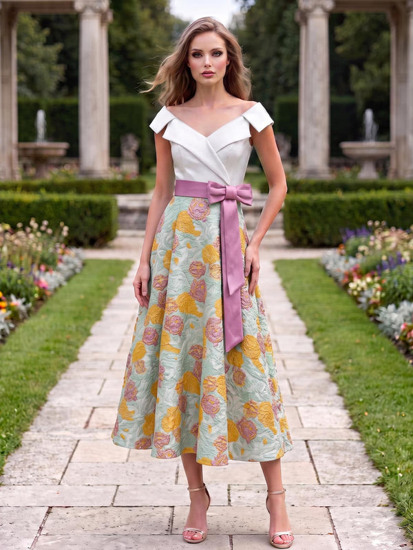 Front view of a floral jacquard satin tea-length mother of the bride dress with an off-the-shoulder fold-over neckline, fitted white bodice, contrast pink bow sash, and full A-line skirt.