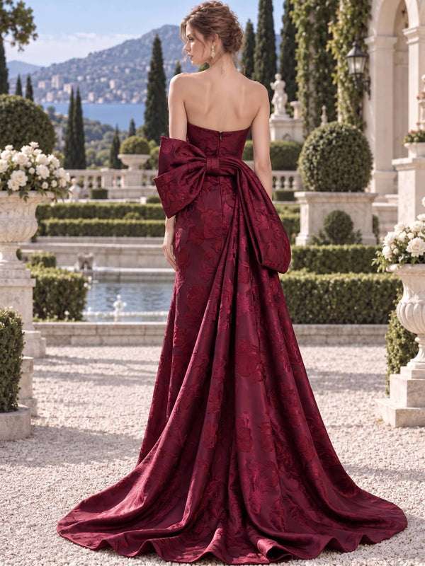 Back view of a burgundy floral jacquard satin mother of the bride gown featuring a strapless bodice, concealed center back zipper, oversized bow, dramatic draped side panel and sweep train.