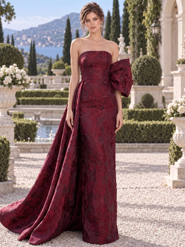 Front view of a burgundy floral jacquard satin mother of the bride gown with strapless neckline, fitted column silhouette, oversized sculptural side bow, draped panel and floor-length hem.