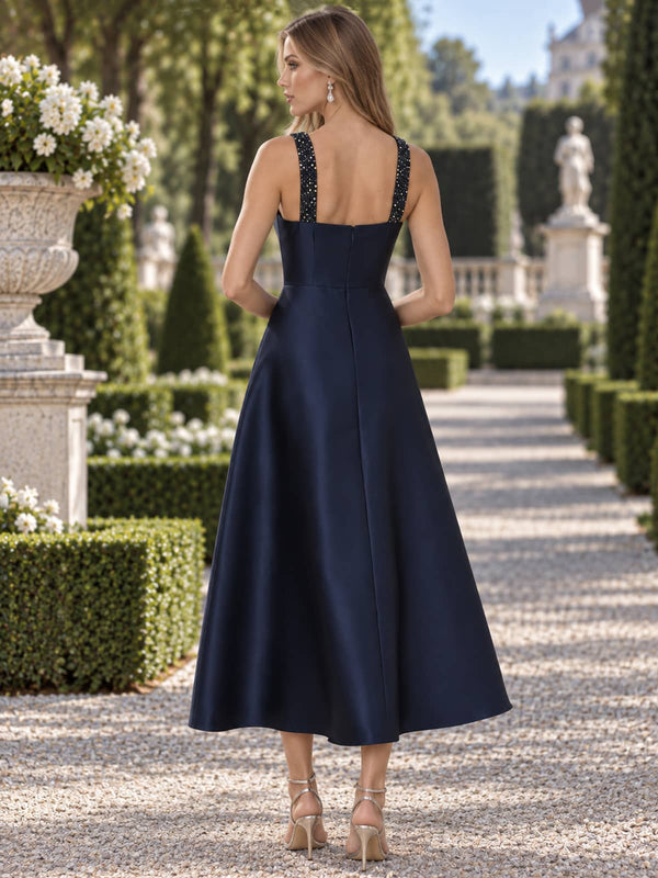 Back view of a navy satin mother of the bride dress featuring embellished shoulder straps, a clean square-inspired back neckline, fitted waist, concealed center-back zipper and tea-length A-line skirt.