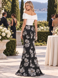 Back view of a black and ivory floral jacquard satin mother of the bride dress featuring an off-the-shoulder ivory draped bodice, center-back closure, fitted hips and oversized silver-gray floral jacquard floor-length mermaid skirt.