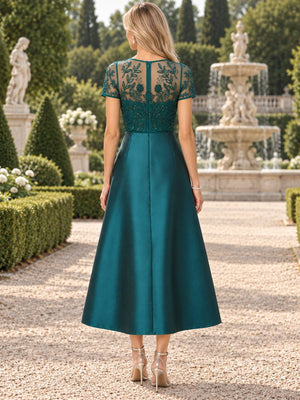 Back view of an emerald green mother of the bride dress featuring a sheer embroidered lace upper back, short sleeves, tonal beadwork, center back zipper and a structured satin A-line tea-length skirt.