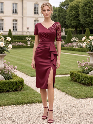 Front view of a burgundy satin mother of the bride dress featuring a wrap-inspired V-neck bodice, asymmetric sheer lace panel with tonal 3D floral appliqués, short sleeves, ruched waist, cascading side ruffle and fitted knee-length sheath skirt with a front slit.